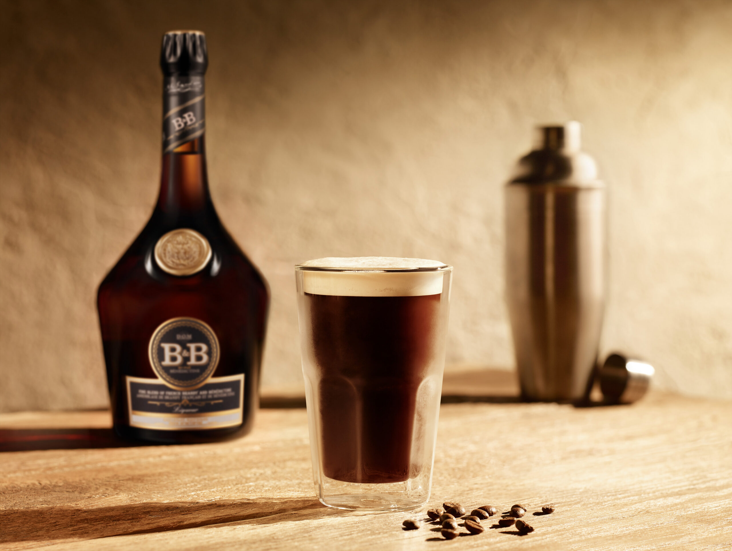 Bouteille de liqueur Bénédictine B&B avec un verre de café noir et un shaker sur une surface en bois.