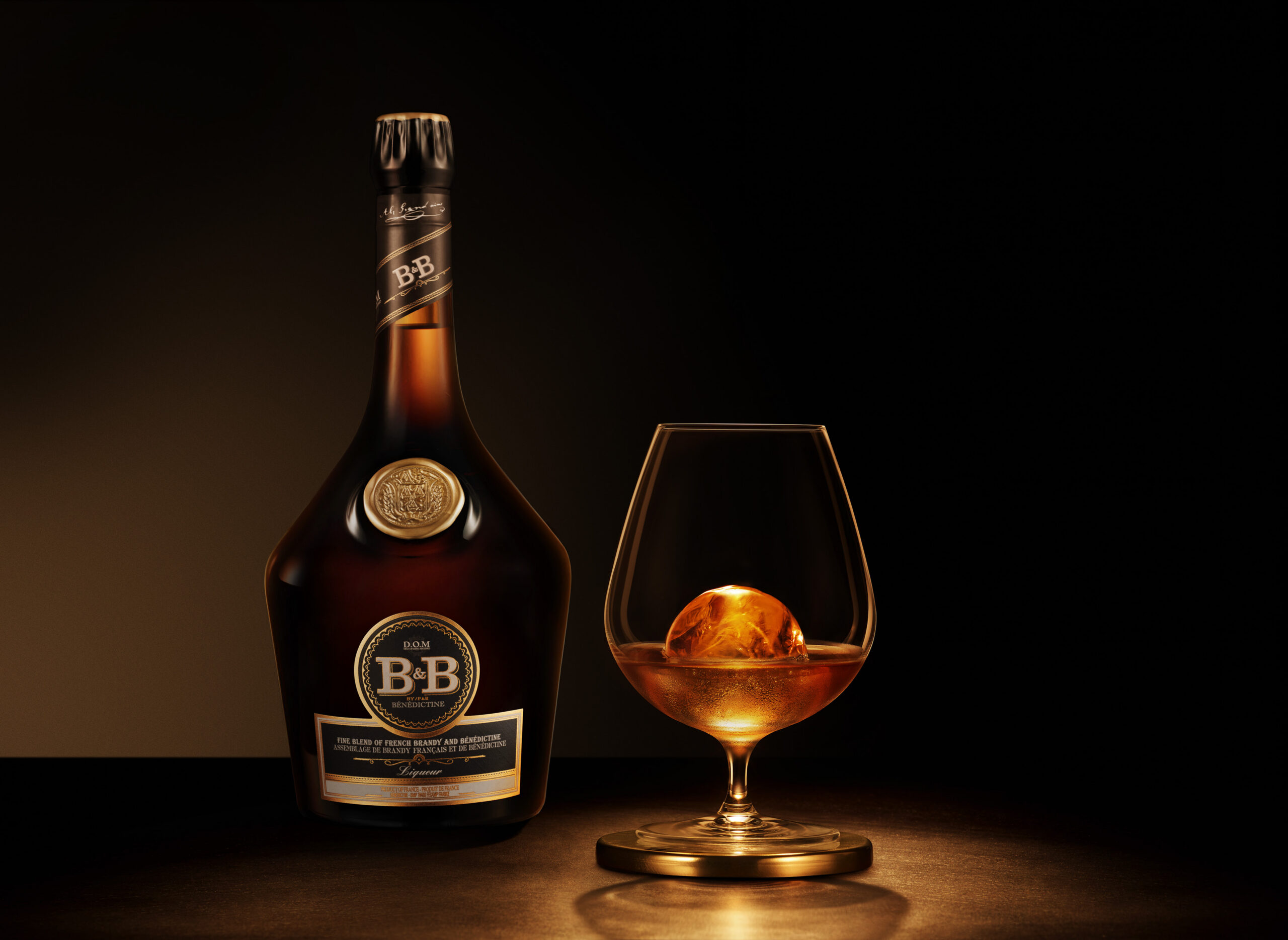 Bouteille de liqueur B&B à côté d'un verre avec la boisson servie sur un gros cube de glace sphérique, sur fond sombre.